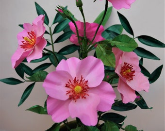 May include: A close-up of a pink paper flower arrangement. The flowers are made of paper and have a realistic appearance. The flowers are arranged in a vase and are surrounded by green leaves.