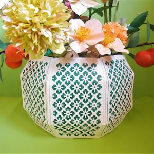 May include: A white ceramic vase with a green floral pattern. The vase is filled with paper flowers, including a yellow chrysanthemum, pink roses, and orange carnations.