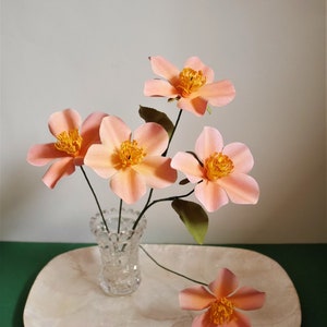 May include: A bouquet of five delicate peach-colored paper flowers with yellow centers arranged in a clear glass vase on a white tray. The flowers are arranged in a simple, elegant style.