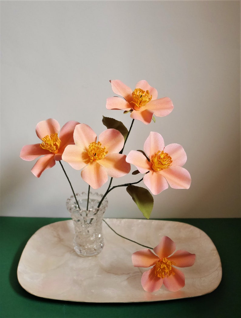 May include: A bouquet of five delicate peach-colored paper flowers with yellow centers arranged in a clear glass vase on a white tray. The flowers are arranged in a simple, elegant style.