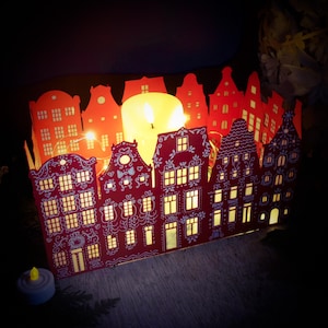 Peut inclure: Un photophore en papier rouge en forme de rangée de maisons, avec des fenêtres éclairées et des découpes décoratives. Une grande bougie à l'intérieur diffuse une lueur chaleureuse. Des petites guirlandes lumineuses et une petite bougie complètent l'ambiance.