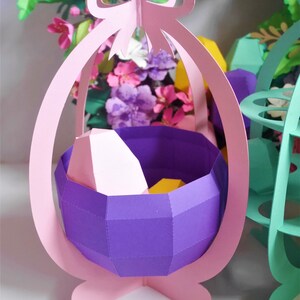 May include: A paper Easter basket in a light pink color, with a purple interior and a bow at the top. The basket is filled with geometric shapes in pink and yellow. The background features colorful paper flowers.