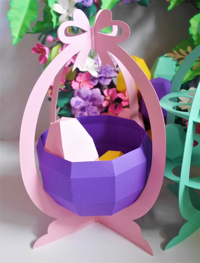 May include: A paper Easter basket in a light pink color, with a purple interior and a bow at the top. The basket is filled with geometric shapes in pink and yellow. The background features colorful paper flowers.