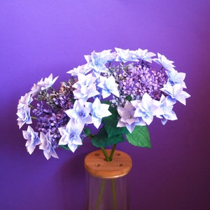 May include: A bouquet of light blue and purple paper hydrangeas in a clear glass vase with a wooden base. The flowers are arranged in a cluster and have a delicate, papery texture.
