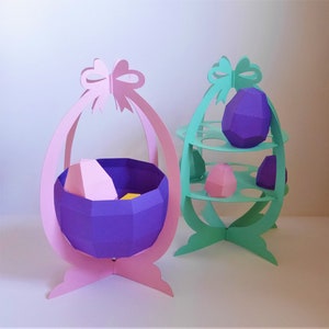 May include: Two Easter egg-shaped paper decorations. One is pink with a purple basket holding a pink and yellow egg. The other is mint green with shelves holding purple and pink paper eggs. Both have bow accents.