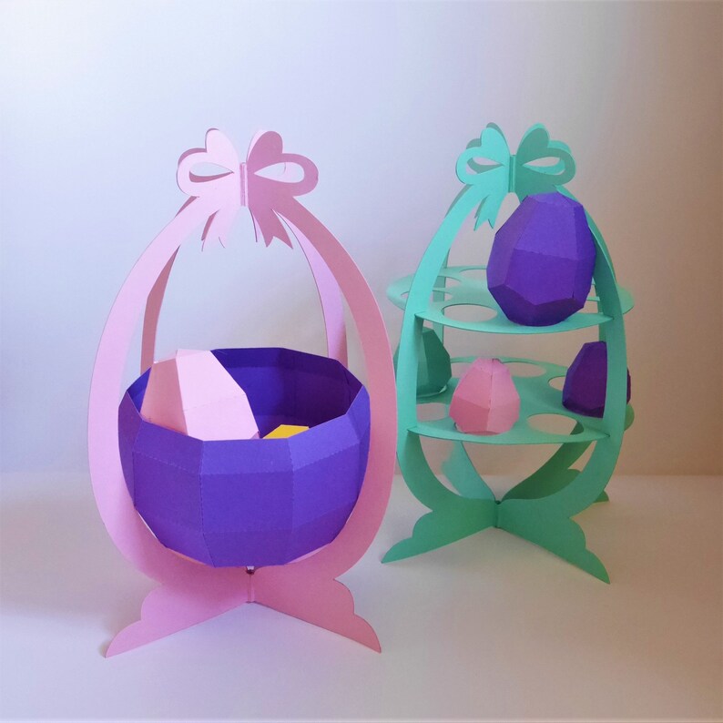 May include: Two Easter egg-shaped paper decorations. One is pink with a purple basket holding a pink and yellow egg. The other is mint green with shelves holding purple and pink paper eggs. Both have bow accents.
