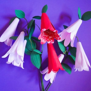 May include: A vibrant arrangement of paper flowers against a purple backdrop. The bouquet features white and red bell-shaped blooms with green leaves. The red flowers have white speckled patterns, creating a striking contrast.