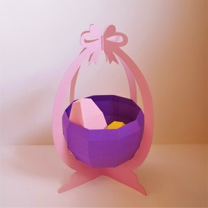 May include: A paper Easter basket in shades of pink and purple. The basket has a bow at the top and a geometric design. Inside the basket are two paper eggs, one pink and one yellow.