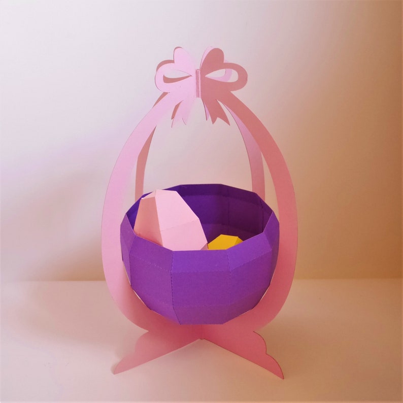 May include: A paper Easter basket in shades of pink and purple. The basket has a bow at the top and a geometric design. Inside the basket are two paper eggs, one pink and one yellow.