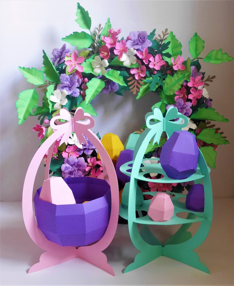 May include: A colorful Easter-themed display featuring paper-crafted egg-shaped baskets and a tiered stand. The display includes a pink basket, a teal stand, and geometric paper eggs in purple, pink, and yellow. A floral wreath with pink and purple flowers is in the background.
