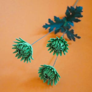 May include: Three green artificial chrysanthemum flowers with detailed petals and stems, and a dark green leaf, against an orange background. The flowers are in various stages of bloom, creating a natural aesthetic.