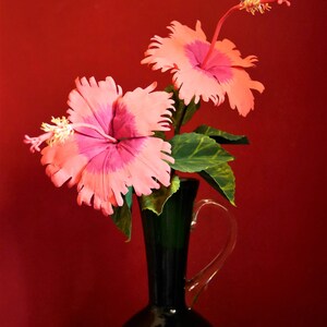 May include: Two pink hibiscus flowers with yellow centers in a dark green vase against a red background.
