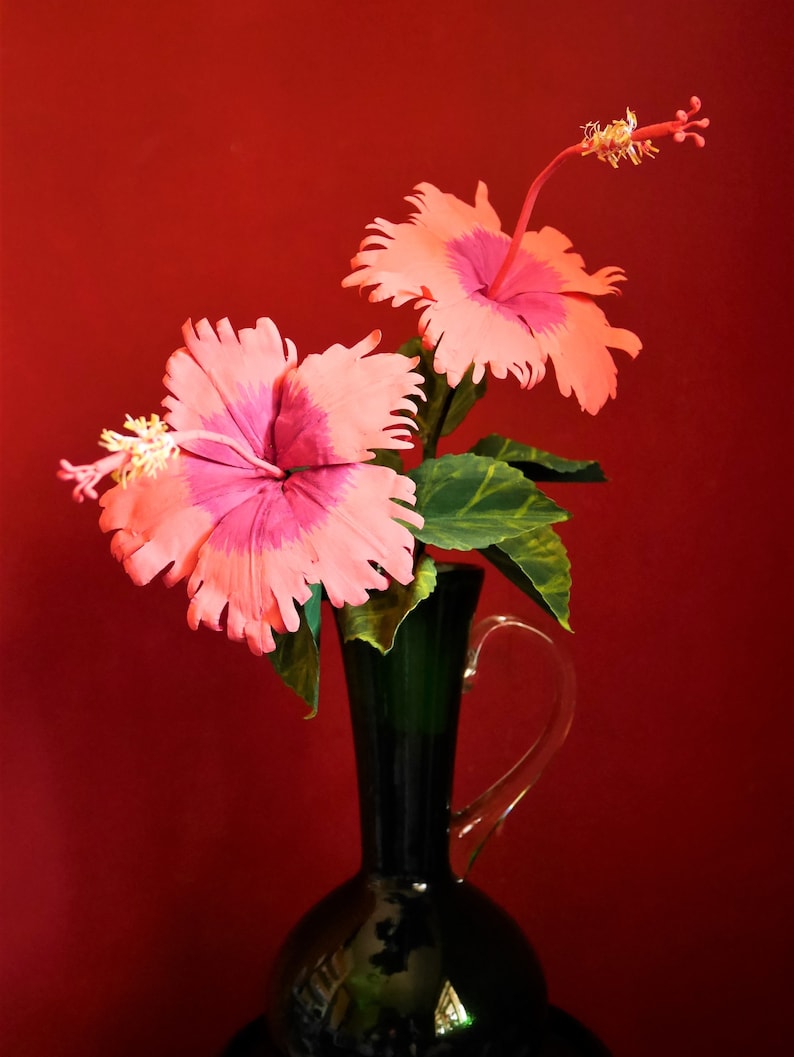 May include: Two pink hibiscus flowers with yellow centers in a dark green vase against a red background.
