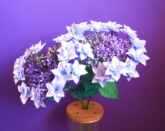 Peut inclure: Un bouquet de fleurs en papier d'hortensias bleu clair et violet dans un vase en verre transparent avec une base en bois. Les fleurs sont disposées en grappe et ont une texture délicate et papier.
