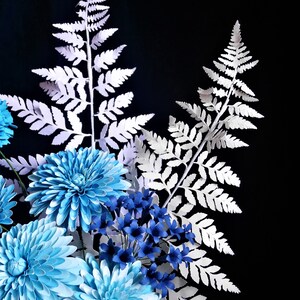 May include: A bouquet of paper flowers in shades of blue and white. The flowers are arranged in a vase and are surrounded by white paper ferns.