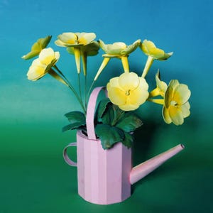 Op de afbeelding: Een roze, geometrische gieter met gele bloemen en groene bladeren. De bloemen hebben een gedetailleerd, realistisch uiterlijk. De gieter heeft een handvat en een tuit, tegen een blauw-groene achtergrond.