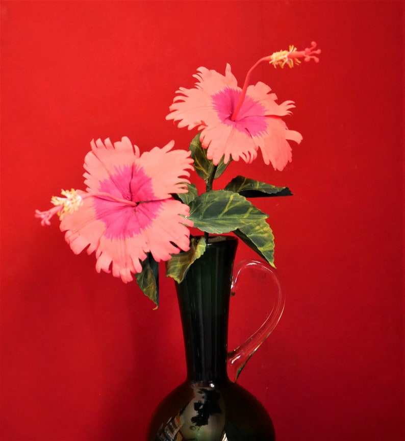 May include: Two pink hibiscus flowers with white centers in a dark green vase against a red background.