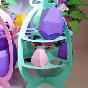 May include: A pastel green paper Easter egg holder with three shelves, holding geometric paper eggs in purple and pink. A matching pink holder is partially visible on the left. A floral wreath is in the background.