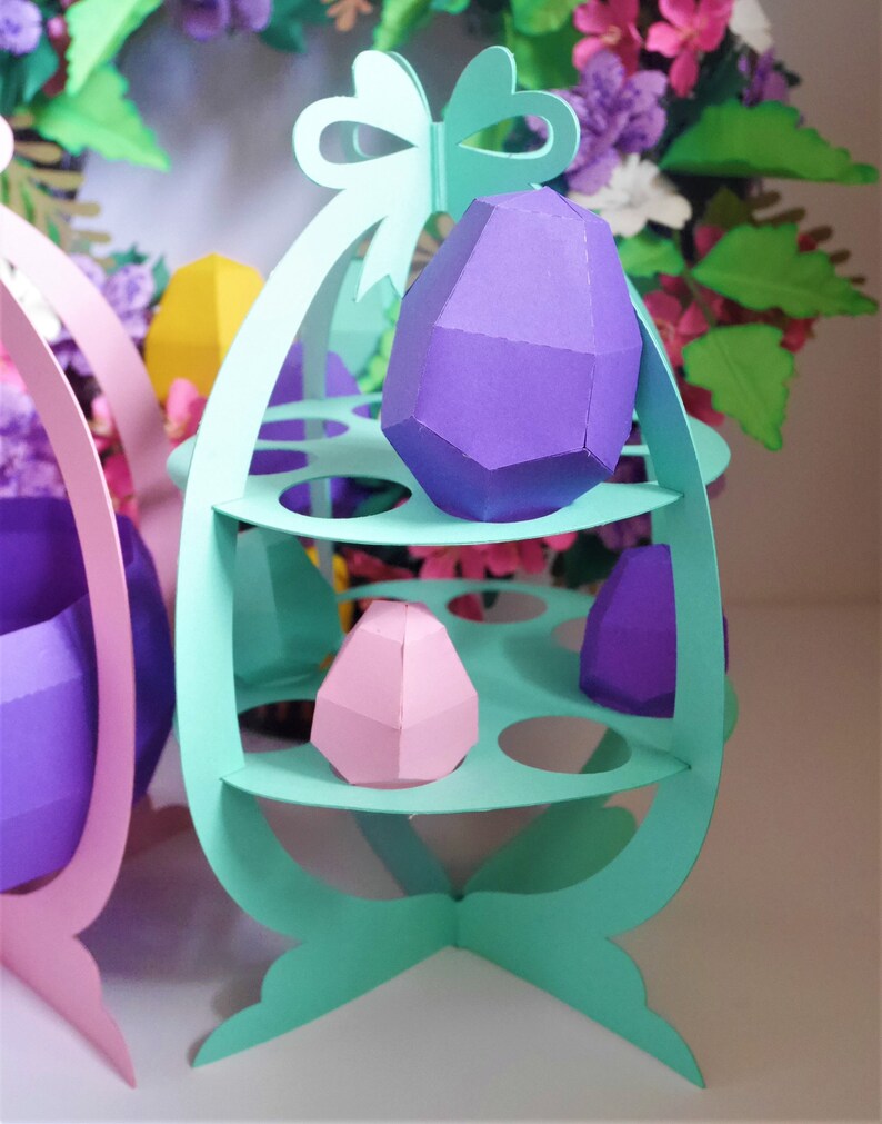 May include: A pastel green paper Easter egg holder with three shelves, holding geometric paper eggs in purple and pink. A matching pink holder is partially visible on the left. A floral wreath is in the background.