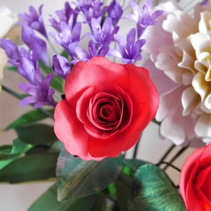 May include: A bouquet of artificial flowers with a red rose, purple flowers, and white flowers. The flowers are arranged in a vase with green leaves.
