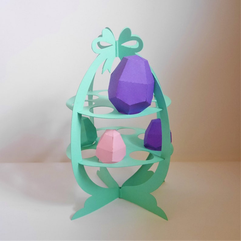 May include: A mint green paper Easter egg display stand with three levels, holding purple and pink geometric paper eggs. The stand has a bow at the top and a decorative base, perfect for Easter decorations.