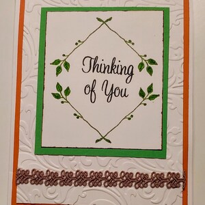 May include: A white card with an orange border and a green frame. The card has a white embossed pattern and a brown lace trim. The text inside the frame reads "Thinking of You" with a green leafy design.