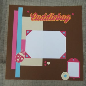 May include: Brown scrapbook page with a white frame, a ladybug, and the text "Cuddlebug". The page is decorated with pink, blue, and beige paper strips and pink and blue hearts.