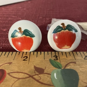 Op de afbeelding: Twee witte keramische knoppen met rode appelmotieven. De appels hebben groene bladeren en stelen. De knoppen zijn ongeveer 2,5 cm in diameter.