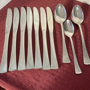 Puede incluir: Un juego de cubiertos de plata con ocho cuchillos y cuatro cucharas. Los cuchillos tienen un mango acanalado y las cucharas tienen un cuenco redondeado.