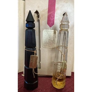 Può includere: Due bottiglie di profumo vintage in una scatola. Una è nera, geometrica e ha un'etichetta dorata con la scritta "Tribute". L'altra è in vetro trasparente con un cordoncino dorato e un liquido dorato all'interno. Un medaglione dorato è appeso a un nastro rosa.