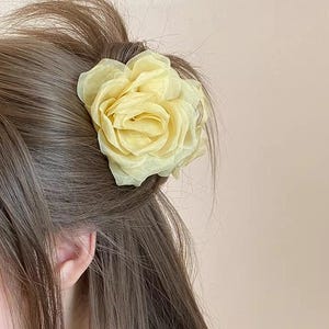 Pode incluir: Uma presilha de cabelo com flor de tecido amarelo pálido, em forma de rosa, está presa em cabelo castanho. A flor, com as suas pétalas delicadas, é o foco principal. O fundo é de uma cor neutra.