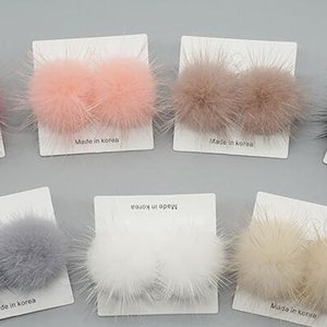 May include: Six pairs of faux fur pom poms on white cards with the text "Made in Korea". The pom poms are in the colors pink, brown, black, gray, white, and beige.