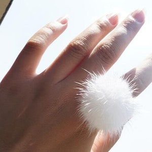 May include: A white fluffy pom-pom ring. The ring is worn on the middle finger of a hand.