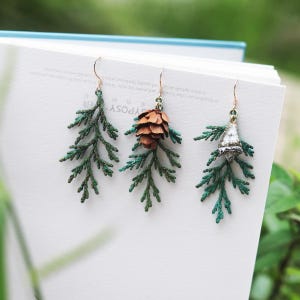 Puede incluir: Tres pares de pendientes colgantes con diseños inspirados en la naturaleza. Cada pendiente presenta una ramita verde y frondosa con un encanto diferente: una piña, una pequeña campana y una ramita simple. Los pendientes se exhiben sobre una superficie blanca.