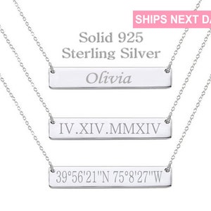 May include: Three silver bar necklaces with different engravings. The top necklace has the name "Olivia" engraved on the bar. The middle necklace has the Roman numerals "IV.XIV.MMXIV" engraved on the bar. The bottom necklace has the coordinates "39°56′21″N 75°8′27″W" engraved on the bar.