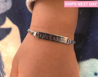 Puede incluir: Una pulsera de plata con una etiqueta rectangular grabada con números romanos. La pulsera está en la muñeca de una persona.