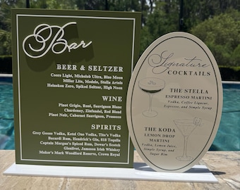 Pode incluir: Uma placa de menu de bar retangular verde-oliva com texto branco listando cervejas, vinhos e destilados, combinada com uma placa oval cor de creme para coquetéis exclusivos com ilustrações de duas taças de martini e ingredientes.