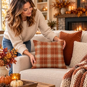 May include: A square throw pillow featuring a rust orange and beige plaid pattern. The pillow is positioned on a light-colored sofa next to a solid rust orange pillow and a textured beige pillow, creating a coordinated set of decorative cushions.