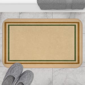 May include: A rectangular bath mat with a light tan center and a border of brown and green stripes. The mat is placed on a tiled floor, with a pair of gray slippers in the foreground. A gray towel is visible in the background.