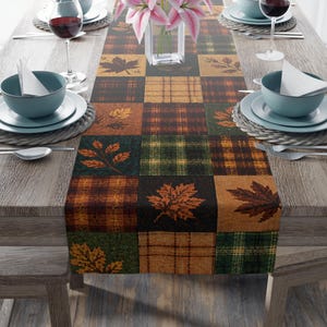 May include: A rectangular table runner featuring a patchwork design of autumn-themed squares. The pattern includes alternating blocks of plaid in shades of orange, brown, green, and yellow, interspersed with detailed illustrations of fallen maple leaves.