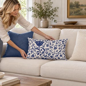 Puede incluir: Un cojín rectangular con un patrón floral azul y blanco de tulipanes y enredaderas estilizadas, colocado sobre un sofá de color claro junto a un cojín cuadrado azul sólido y un cojín cuadrado texturizado de color crema.