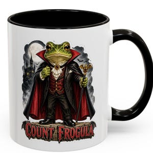 May include: A white ceramic mug with a black handle and rim, featuring a graphic of a green frog dressed as a vampire in a black cape, red waistcoat, and white cravat, holding a bat-topped cane. Below the frog, the text reads Count Frogula.