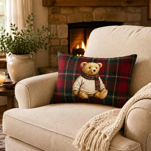 May include: A rectangular throw pillow featuring a dark red, green, and yellow plaid pattern. Centered on the pillow is a graphic of a light brown teddy bear wearing a cream-colored cable knit sweater and a red and green plaid scarf.