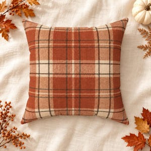 May include: A square throw pillow featuring a plaid pattern in shades of burnt orange, cream, and dark brown. The fabric has a visible woven texture, and the design consists of intersecting horizontal and vertical lines forming a grid.