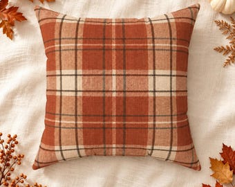 May include: A square throw pillow featuring a plaid pattern in shades of burnt orange, cream, and dark brown. The fabric has a visible woven texture, and the design consists of intersecting horizontal and vertical lines forming a grid.