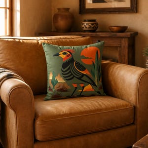 Peut inclure: Un coussin carré décoré d'une illustration graphique représentant un oiseau au plumage orange, jaune et noir sur un fond vert sourd. Le motif comprend un grand soleil orange, des plantes désertiques stylisées et des cactus en aplats.