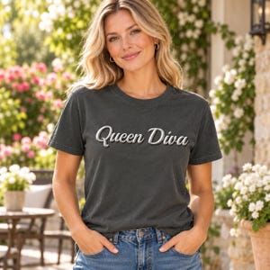 Può includere: Una t-shirt a maniche corte color grigio antracite con la scritta Queen Diva stampata in un carattere corsivo bianco tridimensionale sul petto. Il capo presenta un girocollo e una vestibilità standard, abbinato a jeans in denim blu.