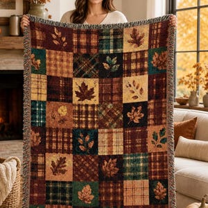 May include: A woven throw blanket featuring a patchwork design of alternating squares. The pattern consists of plaid textures in shades of burgundy, forest green, and gold, interspersed with squares displaying detailed autumn leaf motifs.