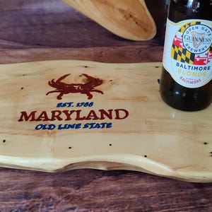 May include: A light-colored wooden serving board with a Maryland crab logo and text. A bottle of Baltimore Blonde beer sits on the board. The board has a natural edge and is on a wooden surface.