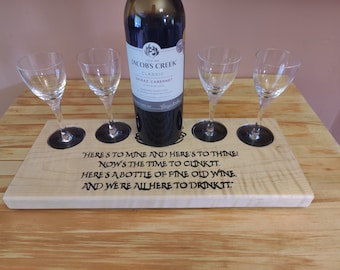 May include: A wooden wine serving tray with four glass holders and a bottle of red wine. The tray has engraved text that reads "Here's to mine and here's to thine! Now's the time to clinkit."